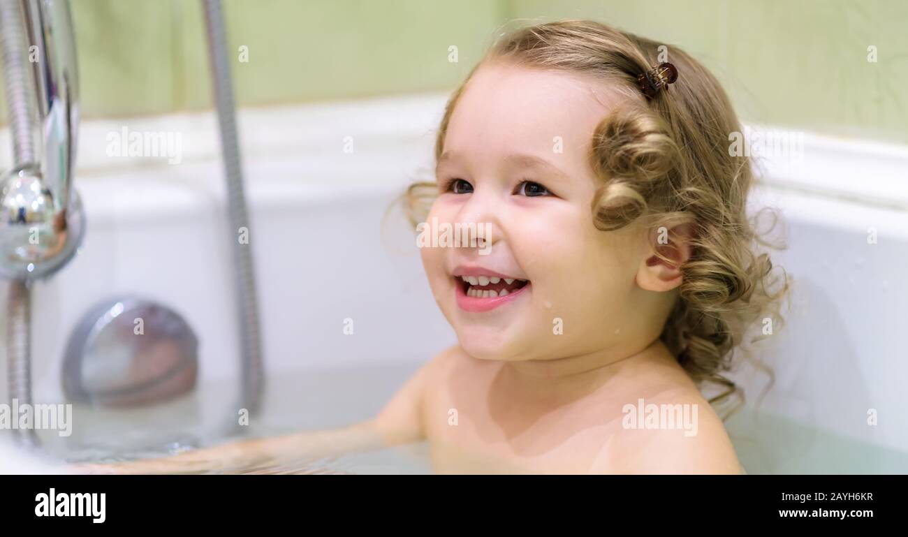 Cheerful baby girl plays in a bath. Beautiful two-year-old child sits in a bathtub and smiles. Adorable toddler while bathing at home. Panoramic view Stock Photo