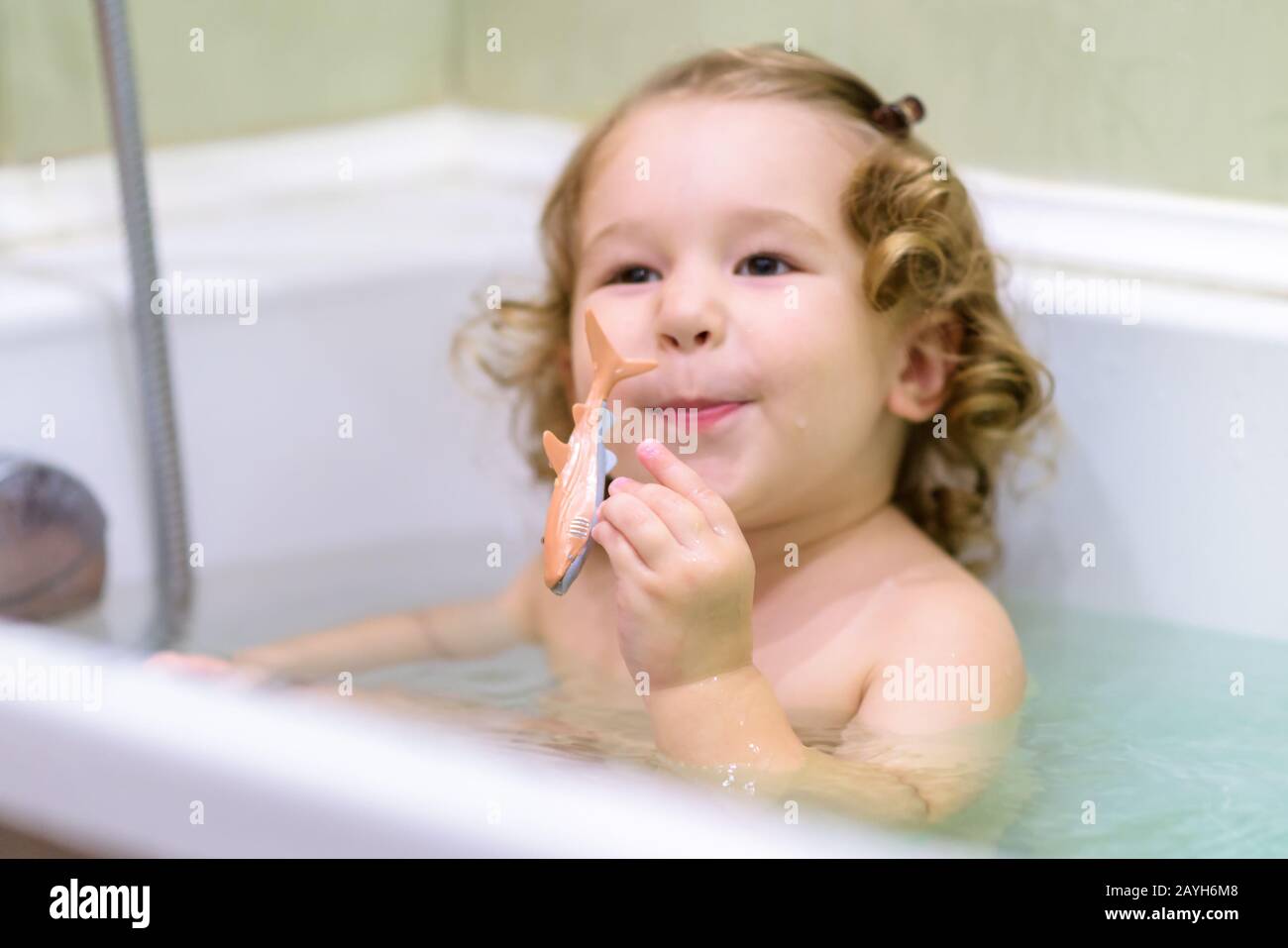 Cheerful baby girl plays in a bath. Two-year-old child holds a toy fish and smiles in a bathtub. Adorable toddler while bathing in the bathroom. Wash Stock Photo
