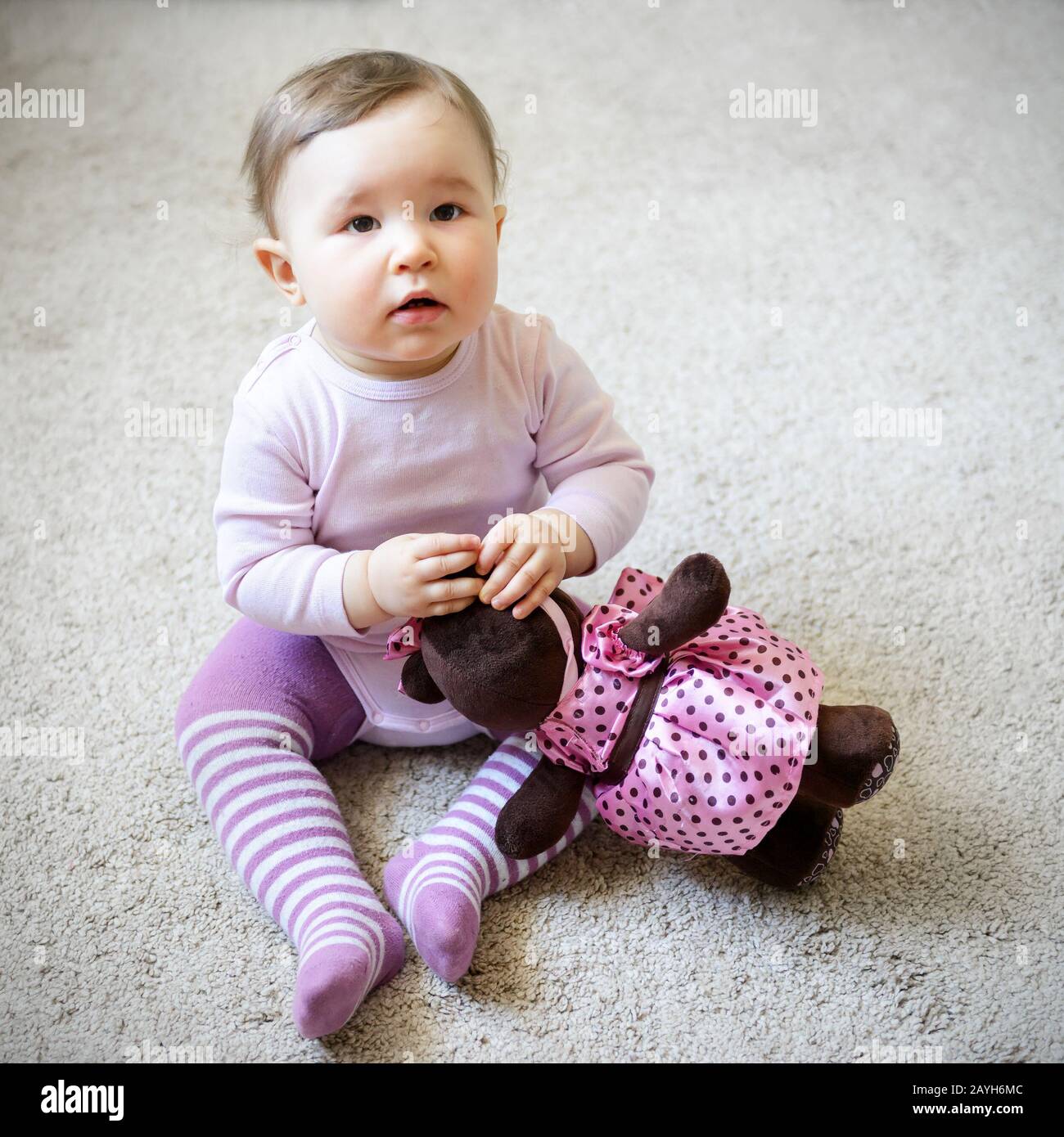 Thoughtful and serious nice baby girl sitting on the floor with toy bear at home Stock Photo
