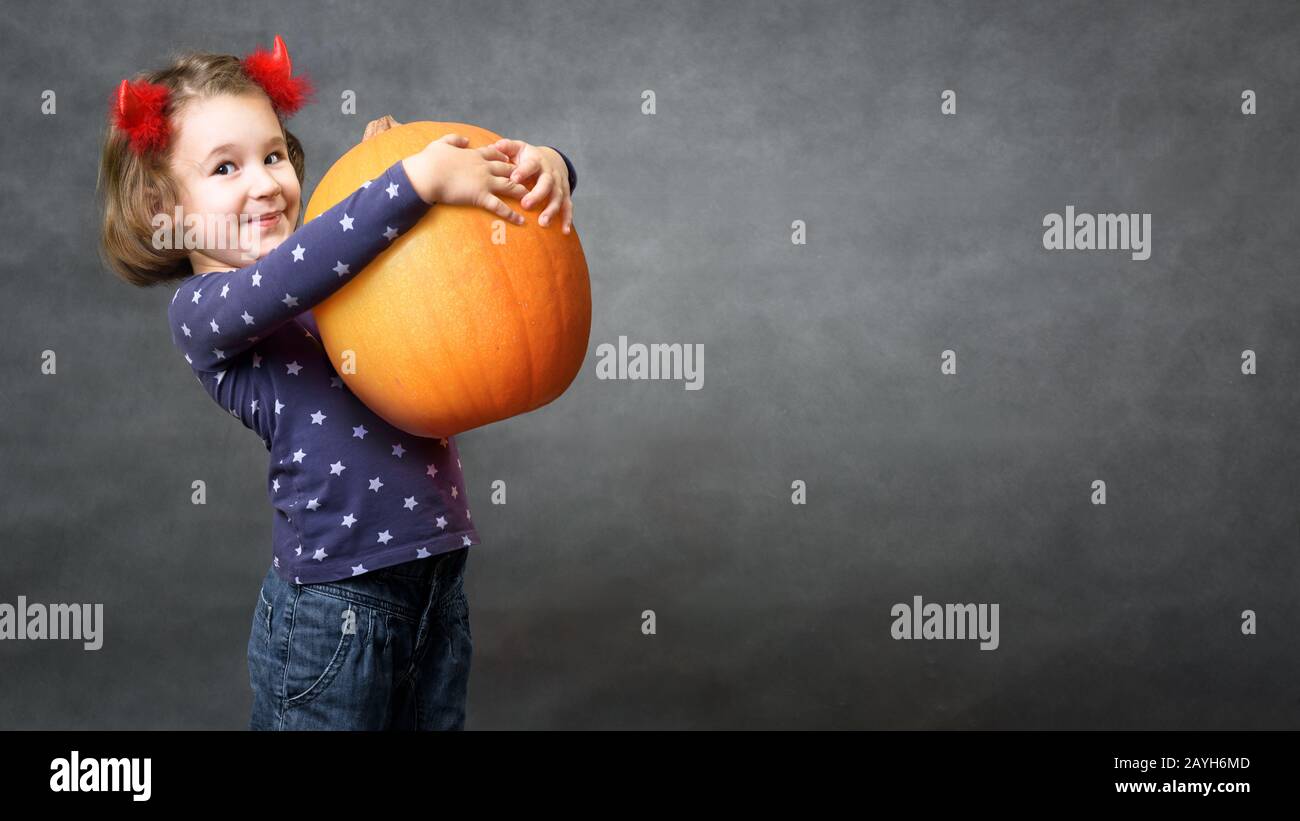 Halloween background with adorable child and space for your text. Little girl with costume horns having fun. Cute toddler holds big pumpkin and smiles Stock Photo