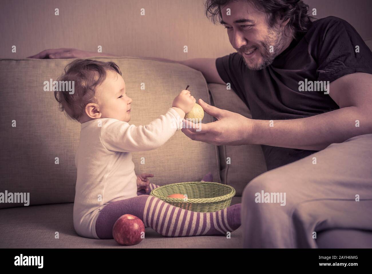 Happy baby girl plays with fruits with her father at home. The one-year child holds a pear in hand. Stock Photo