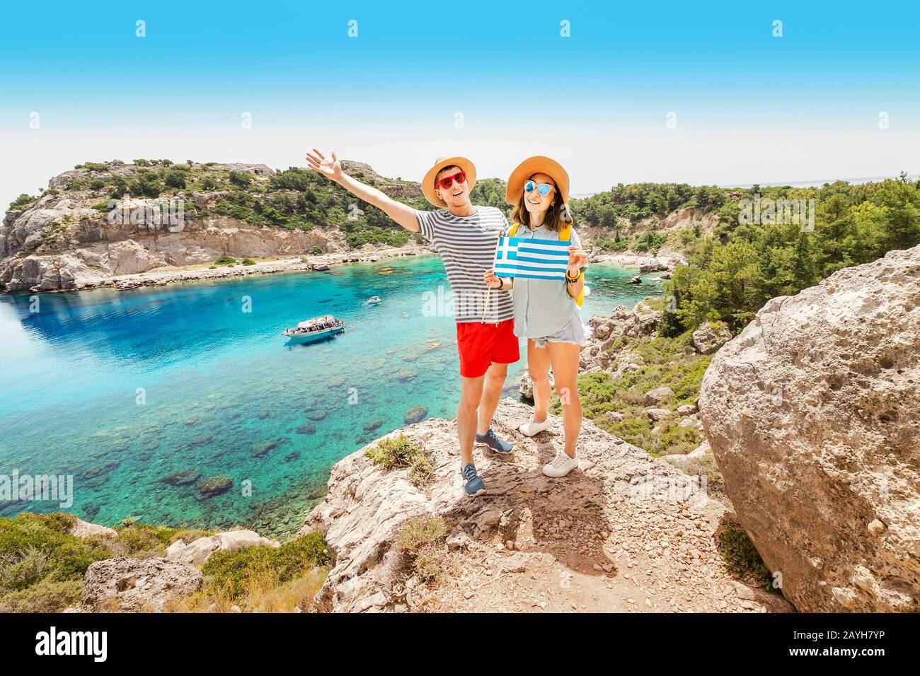 A couple in love hugs and holds a Greek flag, posing against the blue Paradise lagoon. Travel to Greece Stock Photo