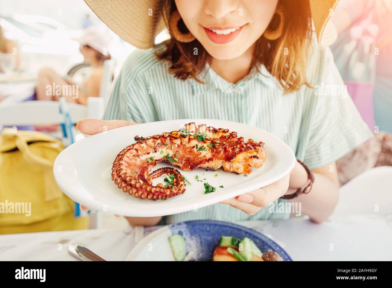 Happy asian woman in hat eating delicious grilled octopus in seafood restaurant. Delicacy and healthy gourmet cuisine Stock Photo