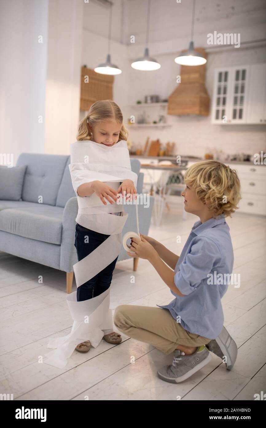 Two kids playing toilet paper mummy game at home and looking involved Stock Photo