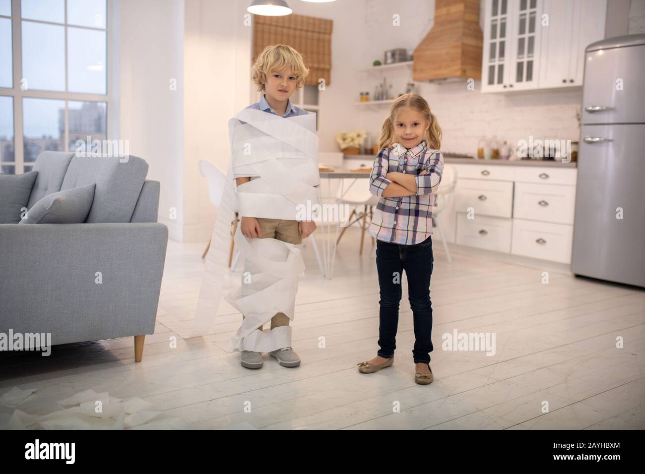 Two kids playing toilet paper mummy game at home and looking amused Stock Photo