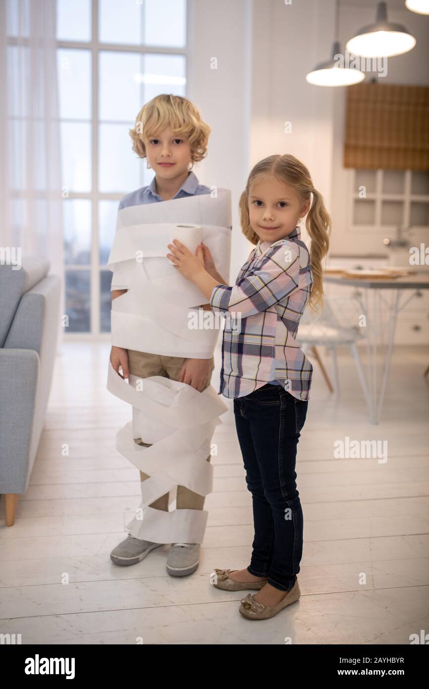 Two kids playing toilet paper mummy game in the kitchen Stock Photo
