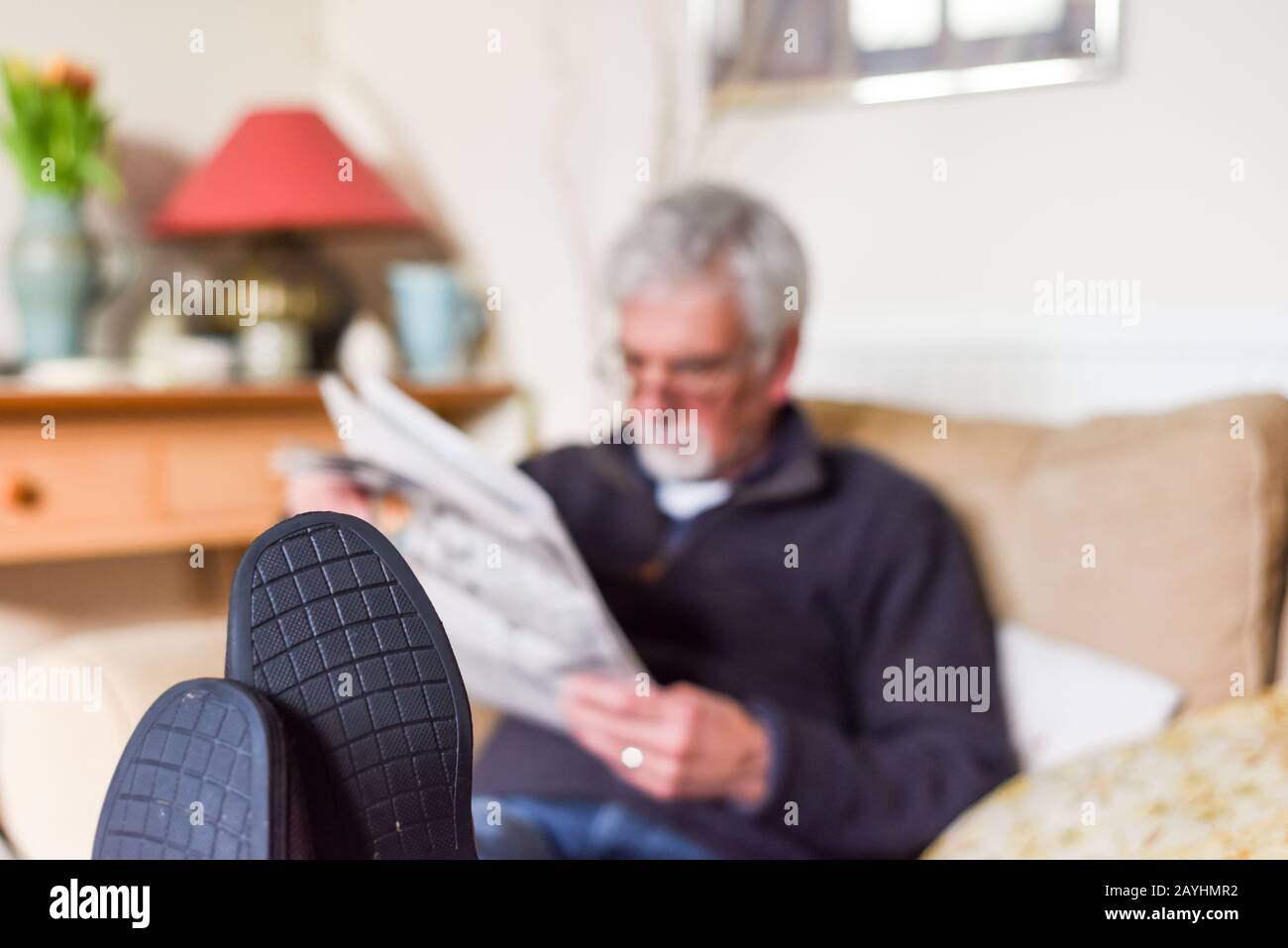 Mature retired man with feet up sitting on a sofa reading a newspaper Stock Photo