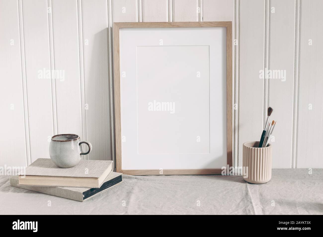 Blank wooden frame mockup with paint brushes, pencils in ceramic holder, cup of coffee and books on linen tablecloth. Artistic scene. Creative table b Stock Photo
