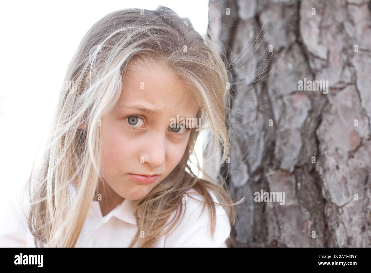 A cute young girl with messed up blond hair and blue eyes, and a half funny, half serious expression on her face, standing next to an old pine tree. W Stock Photo