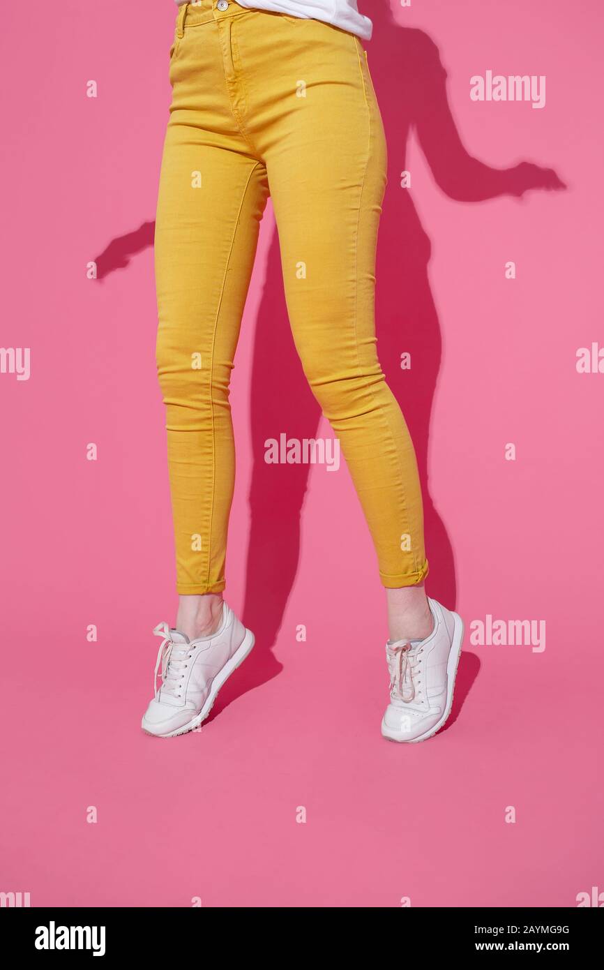 Female feet in trendy yellow jeans and white sneakers on a pink background. Stock Photo