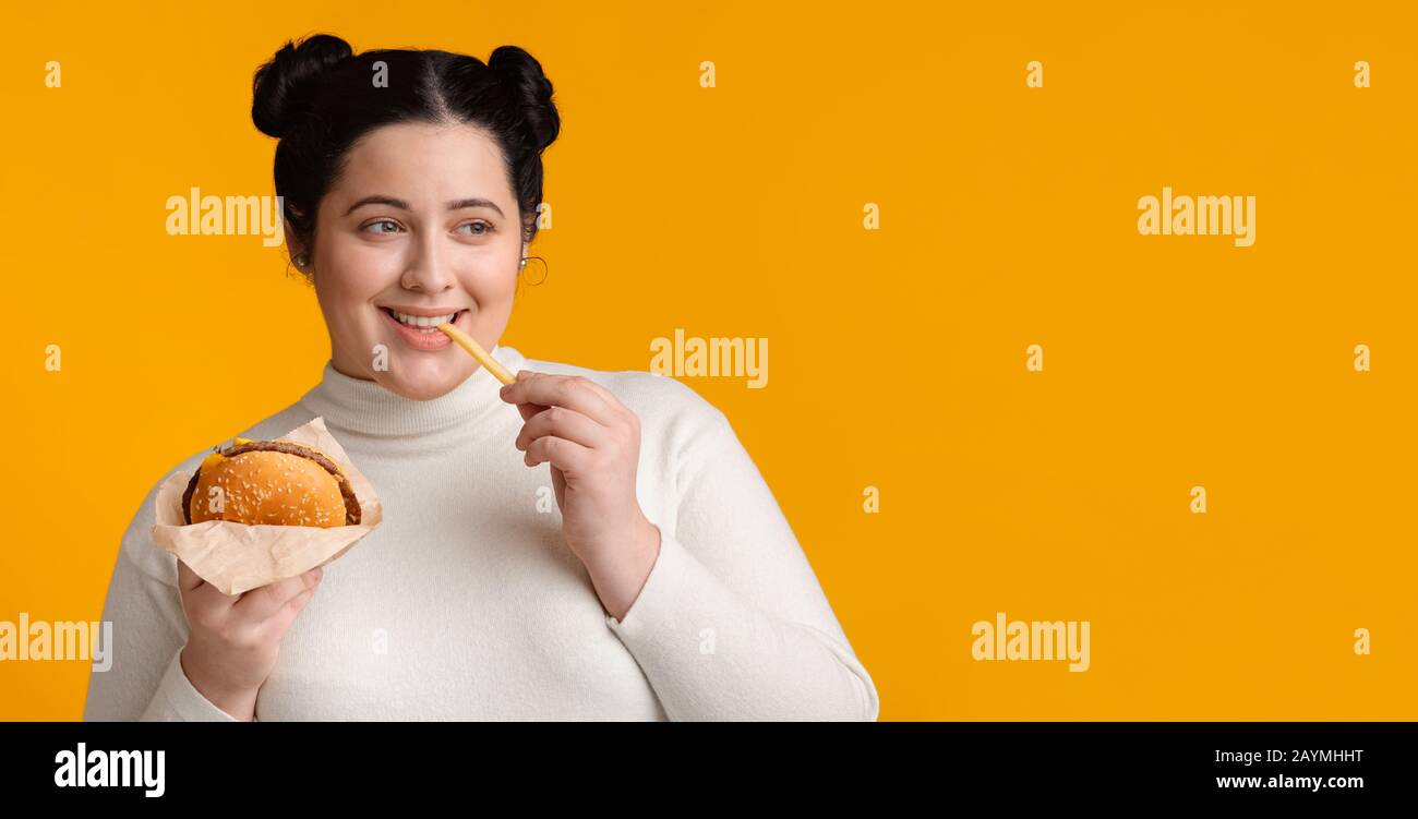 Brunette plump girl eating hamburger and french fries, enjoying fast food Stock Photo