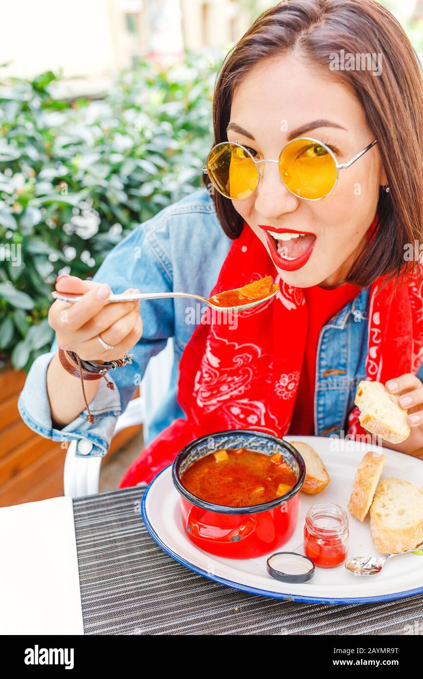 Young beautiful asian girl eating hungarian goulash soup from a decorative red casserole in outdoor restaurant Stock Photo
