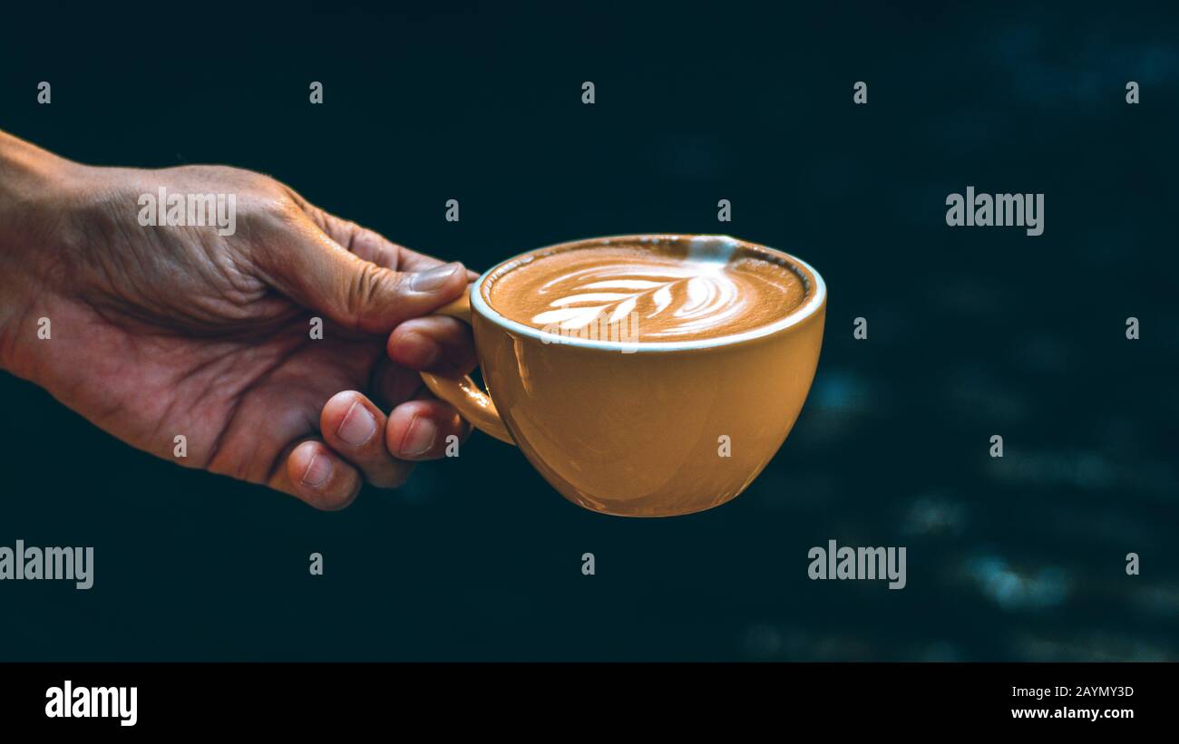 Hand Hold Cup Hot Latte Art in yellow mug against dark background. Stock Photo