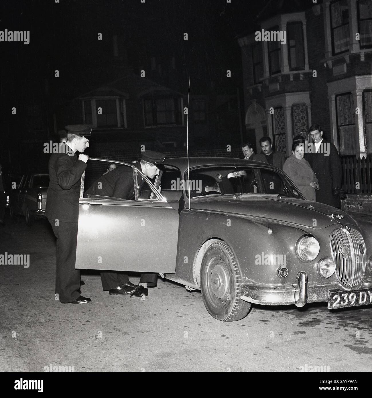 1960s, historical, eveningtime and uniformed policemen inspecting a Jaguar motor car parked in a street in New Cross, South London, England, UK. Stock Photo