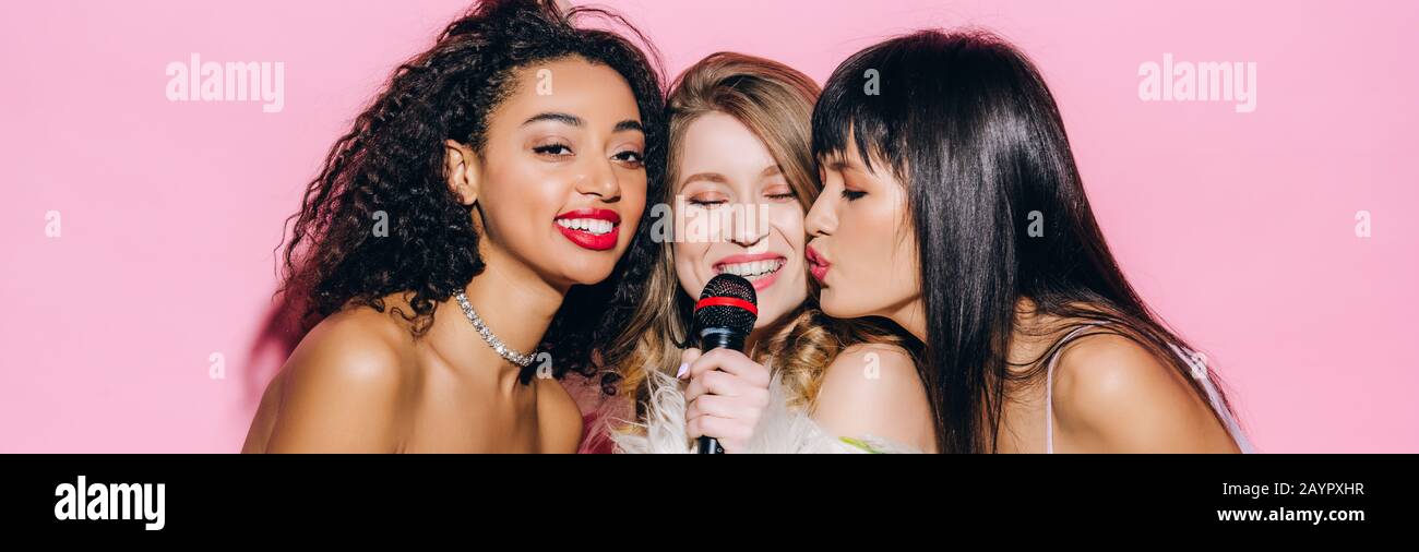 panoramic shot of cheerful multicultural girls singing karaoke with microphone, isolated on pink Stock Photo