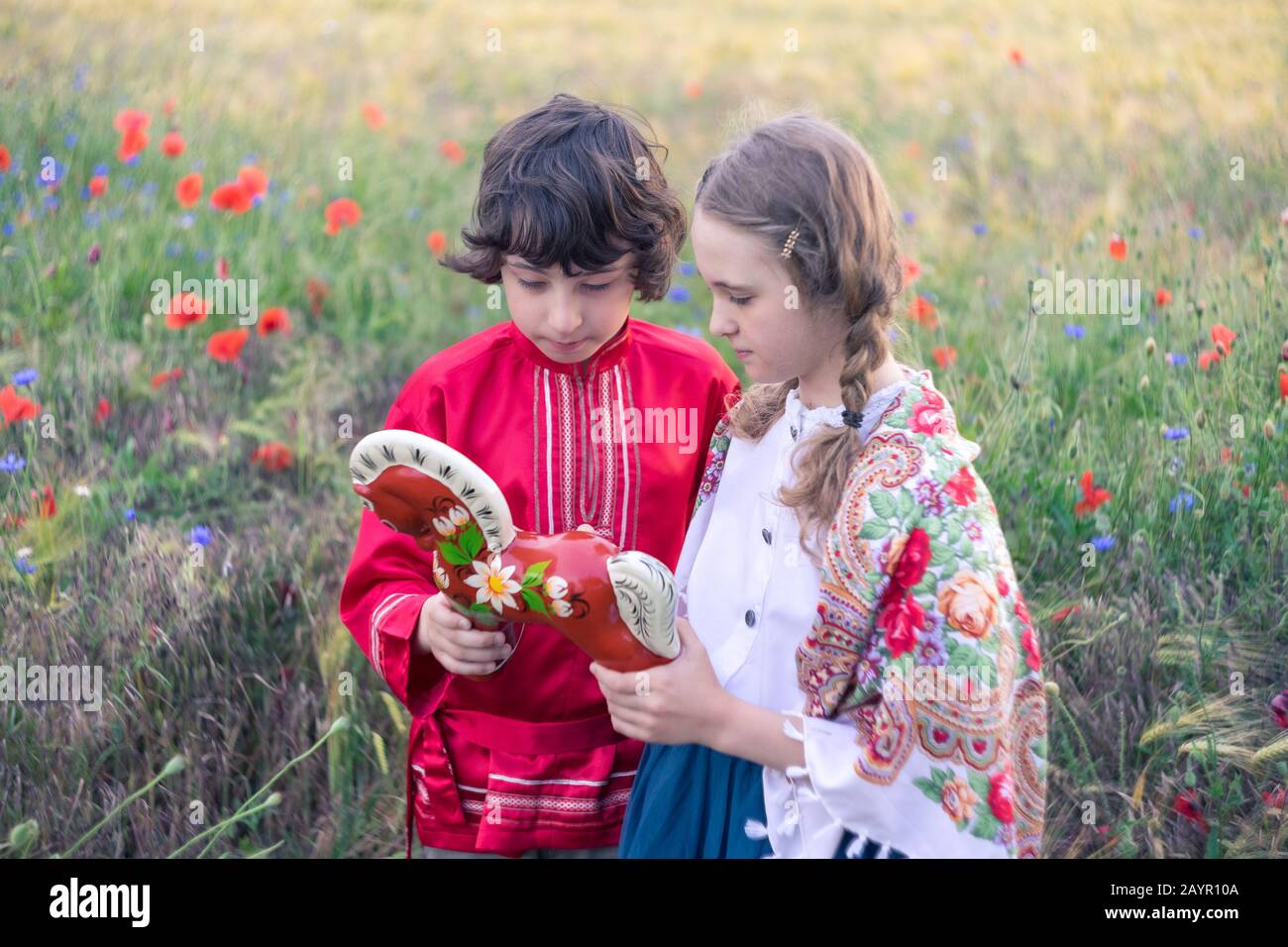 Portrait of a boy and a girl in the field. On the girl is a shawl with folk patterns. Boy in Russian peasant shirt. They are considering a wooden hors Stock Photo