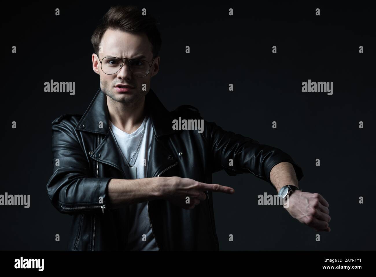 angry stylish brutal man in biker jacket pointing with finger at wristwatch isolated on black Stock Photo