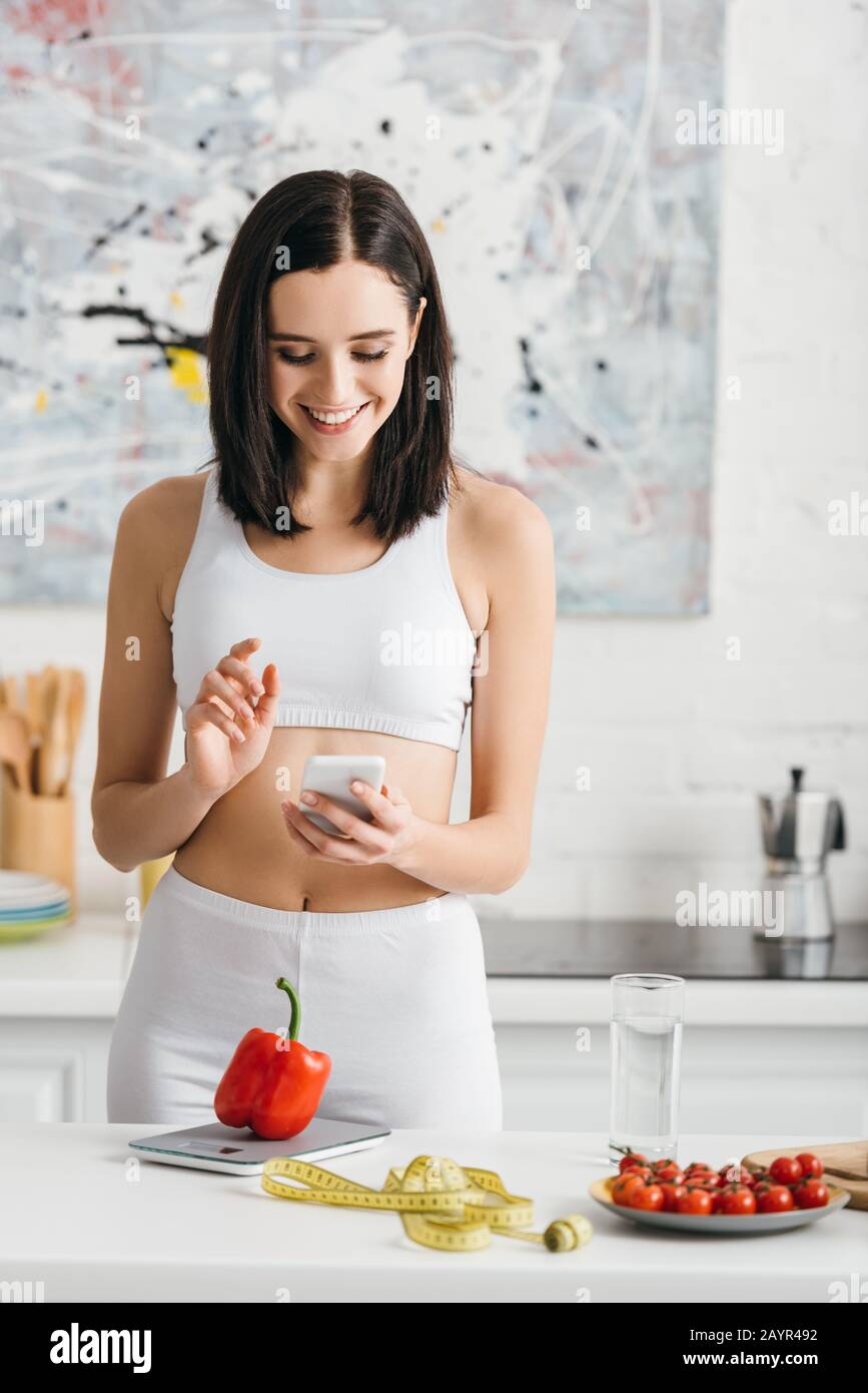 Smiling sportswoman using smartphone near measuring tape, vegetables and scales on kitchen table, calorie counting diet Stock Photo