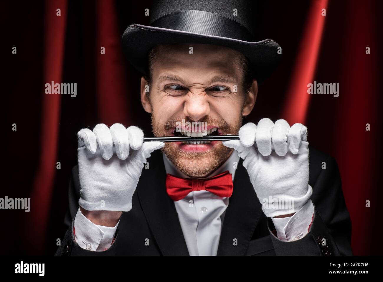 emotional scary magician in suit and hat holding wand in teeth in circus with red curtains Stock Photo