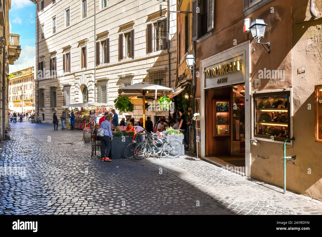Tourists enjoy shopping and lunch at a sidewalk cafe on a narrow cobblestone street in the historic center of Rome, Italy Stock Photo