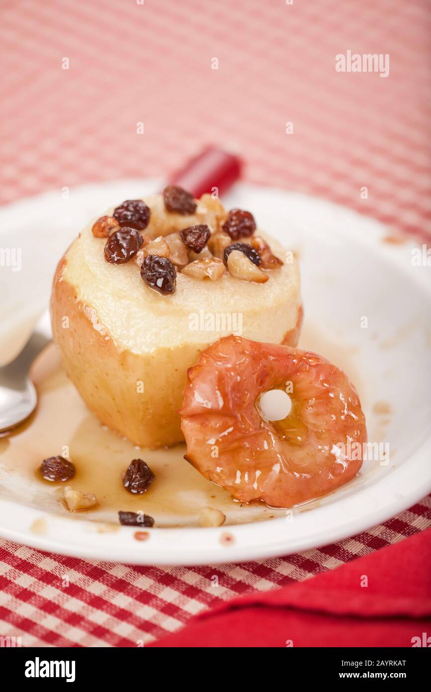 Honey baked apple with the core removed and filled with raisens and walnuts, and drizzled with honey, resting on a white ceramic plate with a red clot Stock Photo