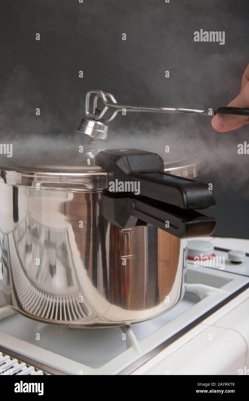 Pressure cooker on a stove with steam escaping as a woman uses tongs to remove the top to let the steam escape Stock Photo