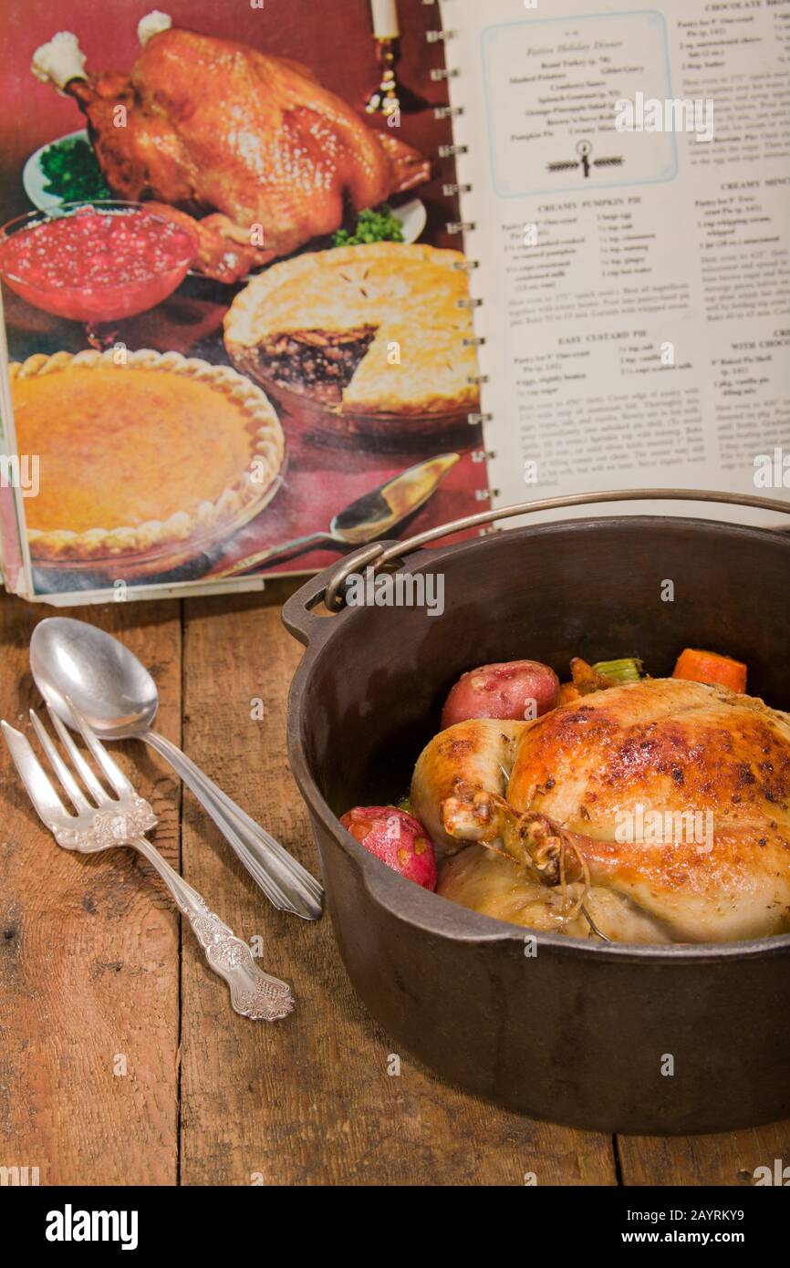 Dutch-style roasted chicken with baked red potatoes, carrots and celery, in a cast iron pot on a rustic wood table, with an old cookbook in the backgr Stock Photo
