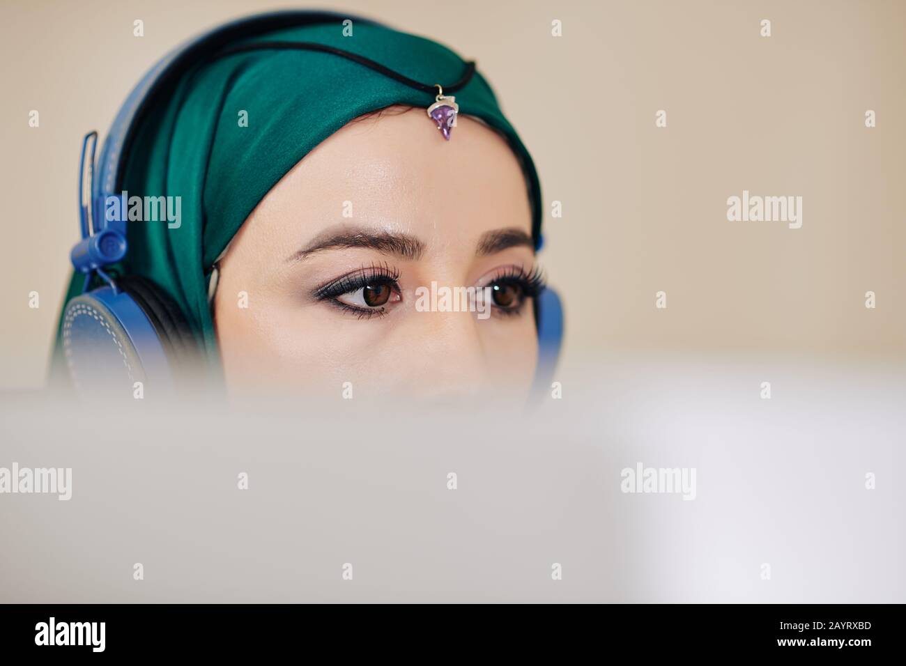 Young beautiful software engineer in hijab listening to music in headphones when programming or editing video on computer Stock Photo