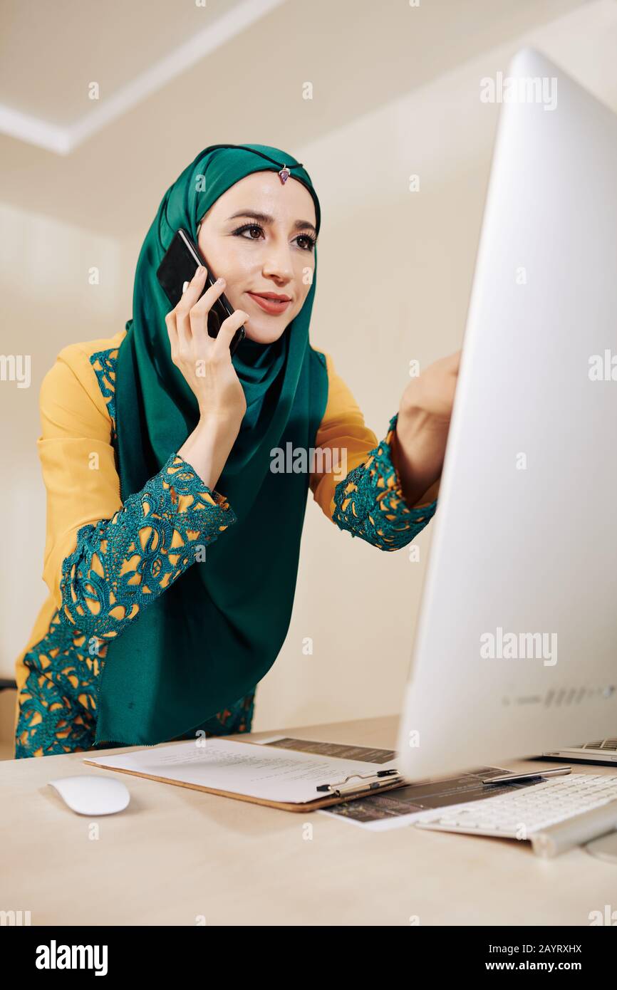 Smiling pretty female engineer talking on phone when discussing strings of code on somputer screen Stock Photo