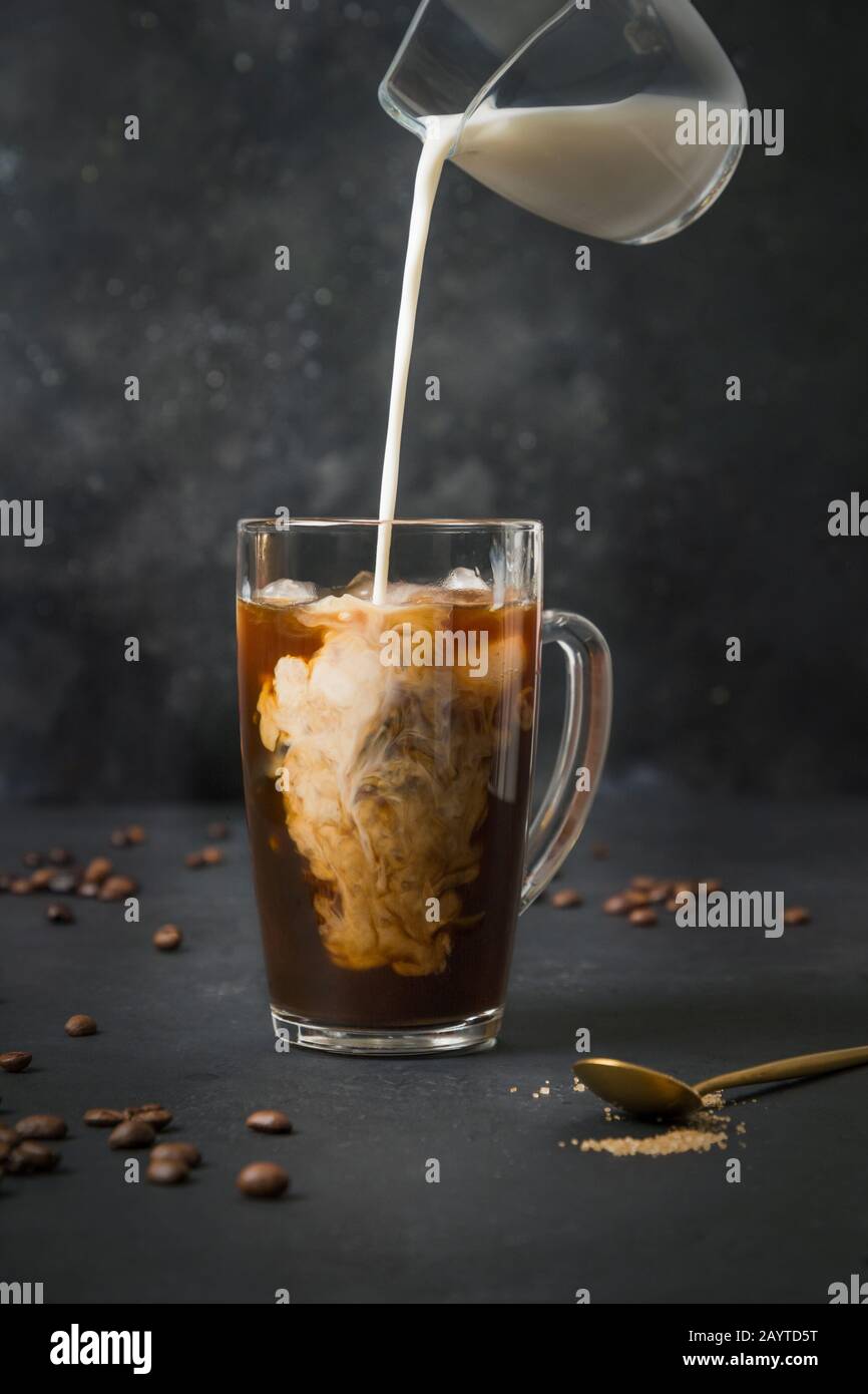 Iced latte coffee in cup glass with pouring milk on black. Close up. Stock Photo