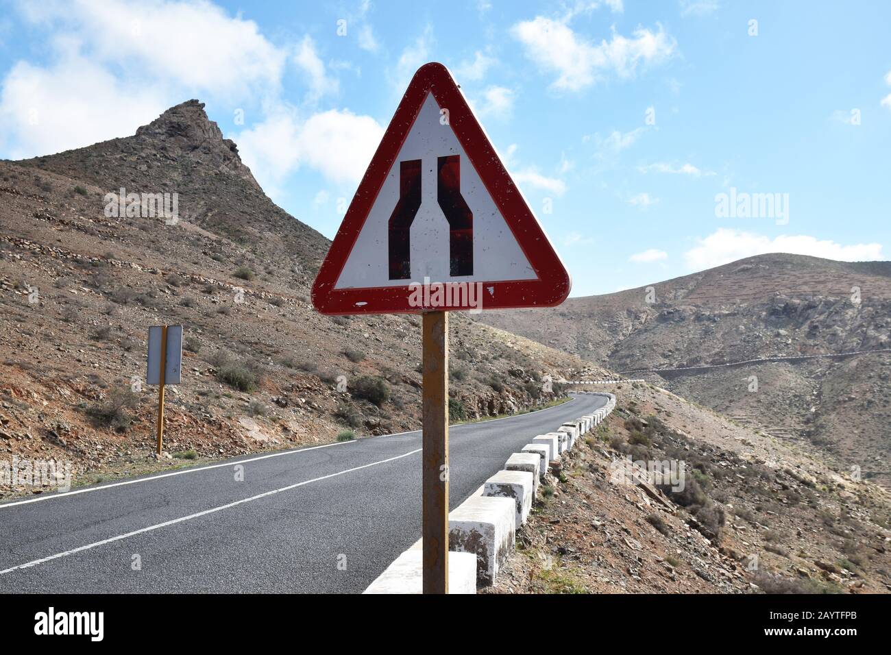 Triangular road narrowing sign on mountain road with mountain in background. Road clearly goes from two lanes to single track. Stock Photo