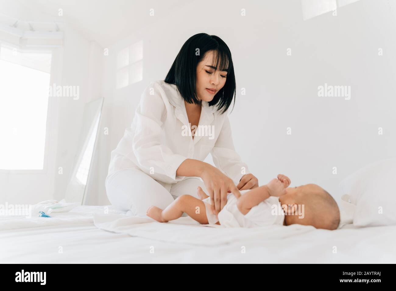 Young Asian mother with closed eyes in white wear changing diaper for