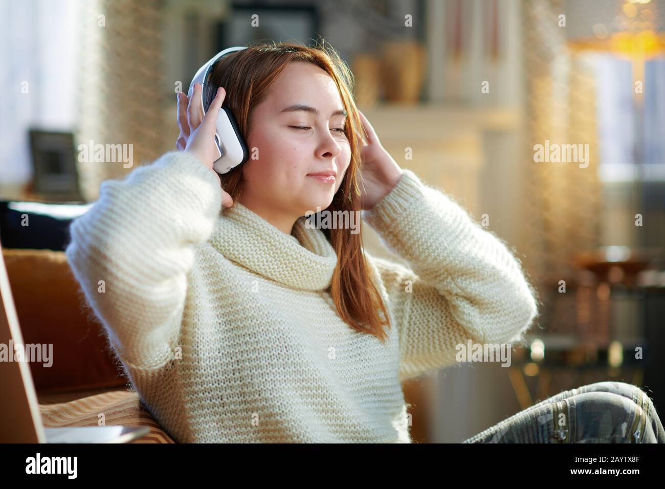 relaxed modern teenager girl with red hair in white sweater listening to the music with headphones while sitting near couch in the modern living room Stock Photo