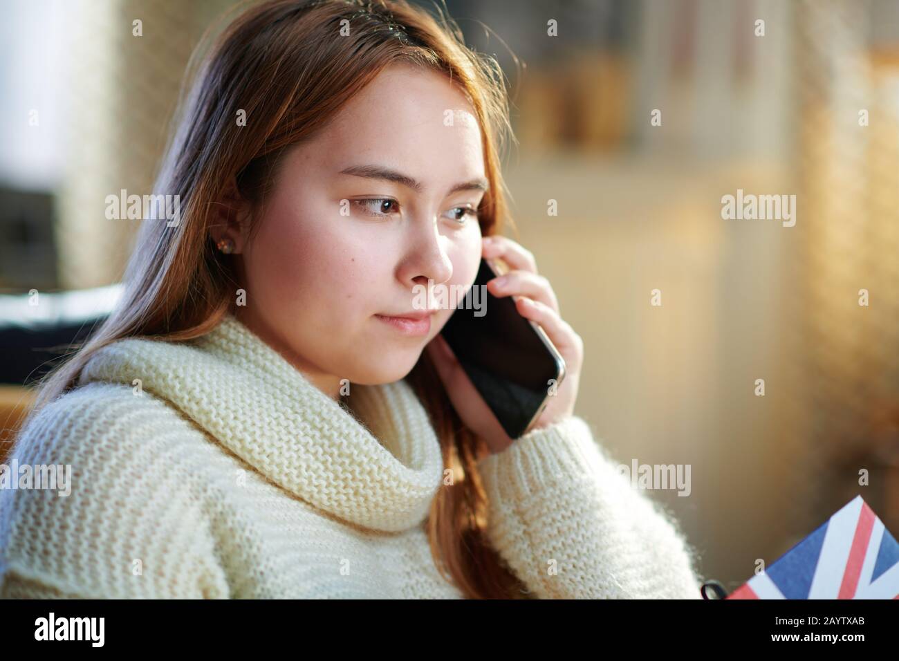 modern teenager girl with red hair in white sweater at modern home in sunny winter day speaking on a cell phone. Stock Photo