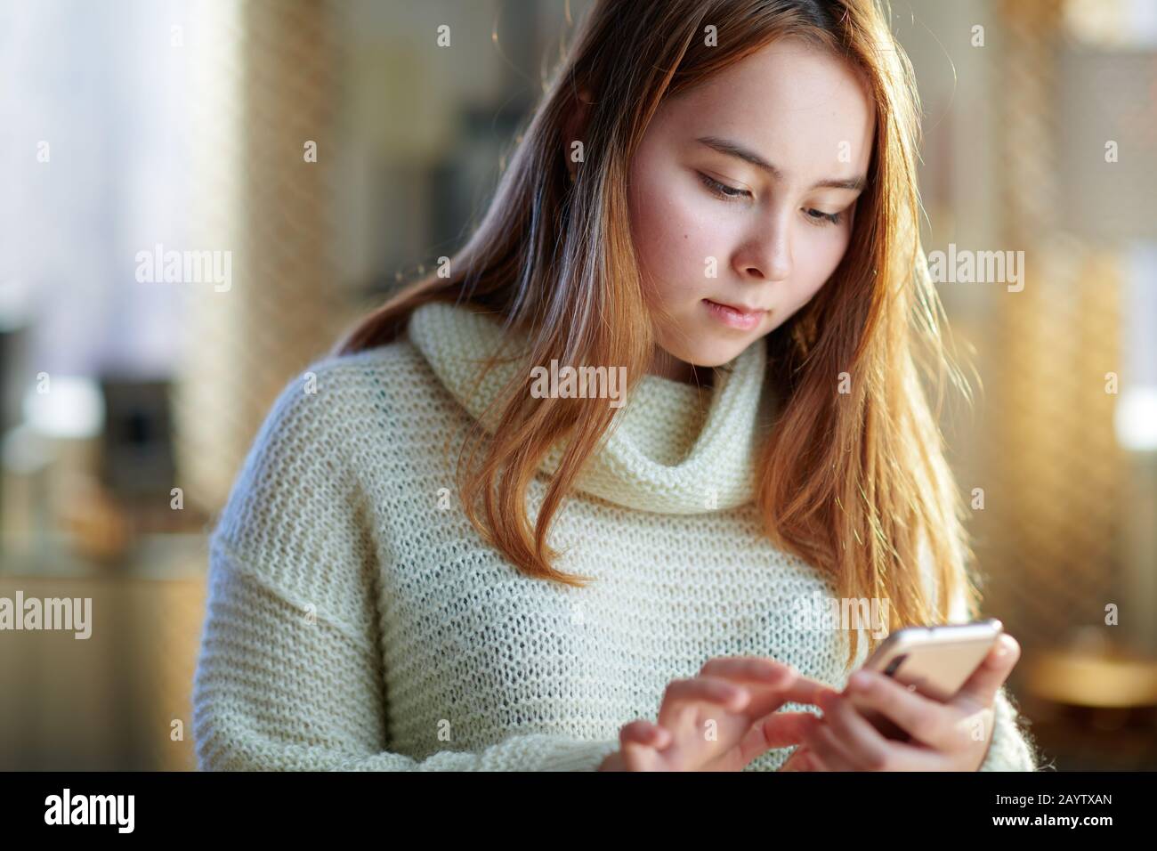 pensive modern teenager girl with red hair in white sweater with smartphone hanging in social media in the modern living room in sunny winter day. Stock Photo