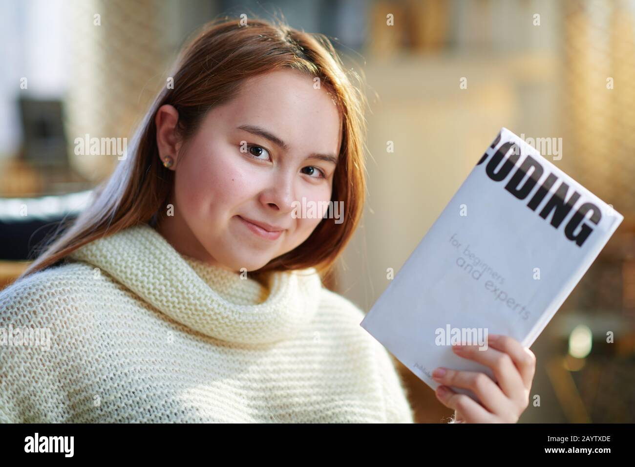 smiling modern teenage girl with red hair in white sweater in the modern house in sunny winter day showing educational coding book. Stock Photo