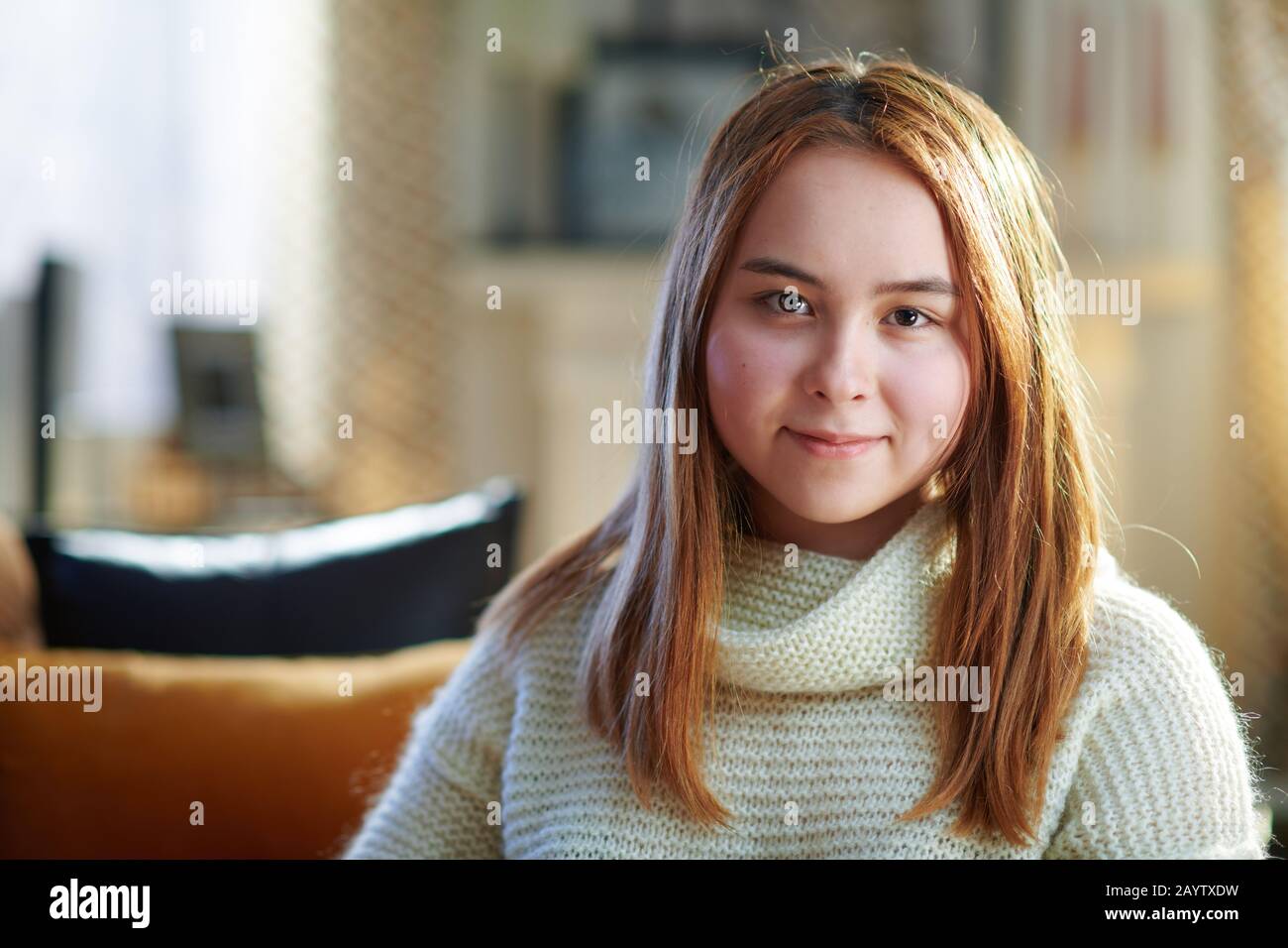 Portrait of happy modern teenager girl with red hair in white sweater at modern home in sunny winter day. Stock Photo