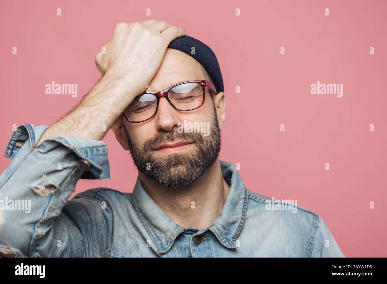 Portrait of depressed middle aged unshaven male closes eyes and keeps hand on forehead, has unhappy expression, isolated over pink background. Fatigue Stock Photo