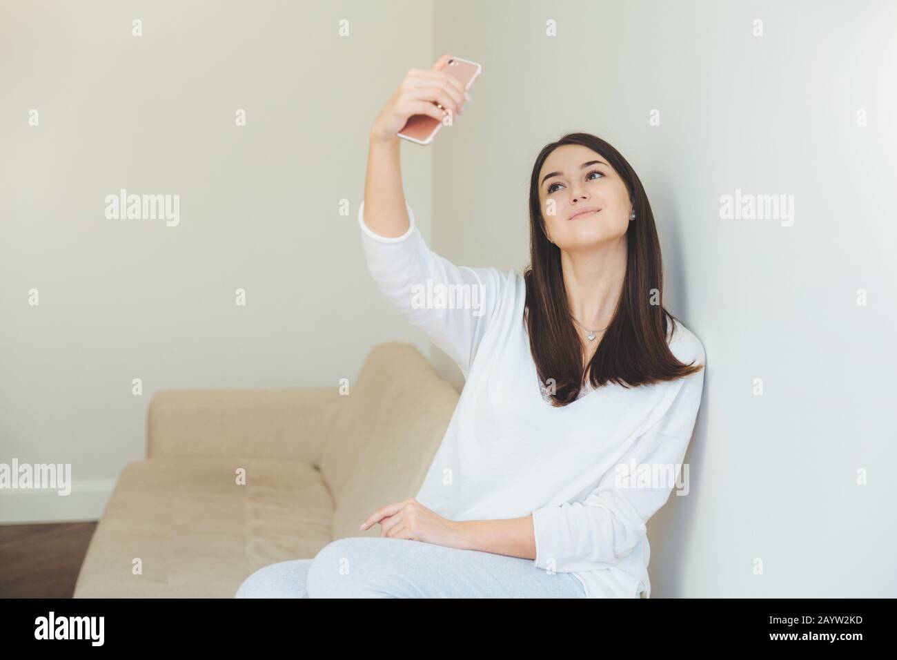 Beautiful woman dressed casually, spends free time at home alone, poses at camera of smart phone, makes selfie, likes to be photographed. Attractive f Stock Photo