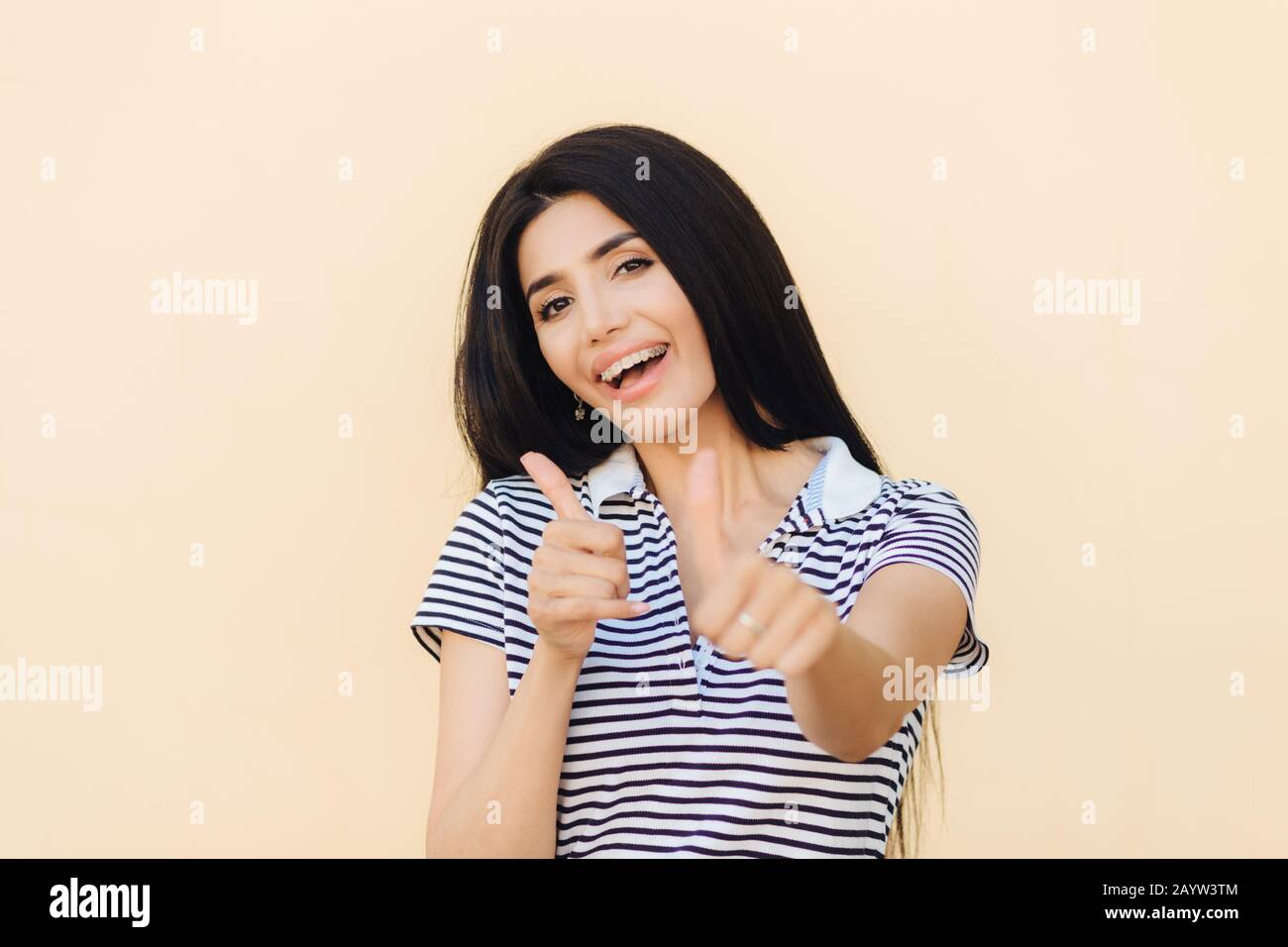 Shot of attractive European female gives double thumb up, has positive expression, has white teeth with brackets, dressed casually, makes okay and app Stock Photo