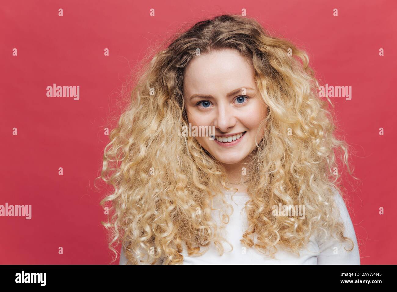 Beautiful blue eyed young female model with gentle smiles, being glad to have date with boyfriend, wears white sweater, isolated over bright pink stud Stock Photo