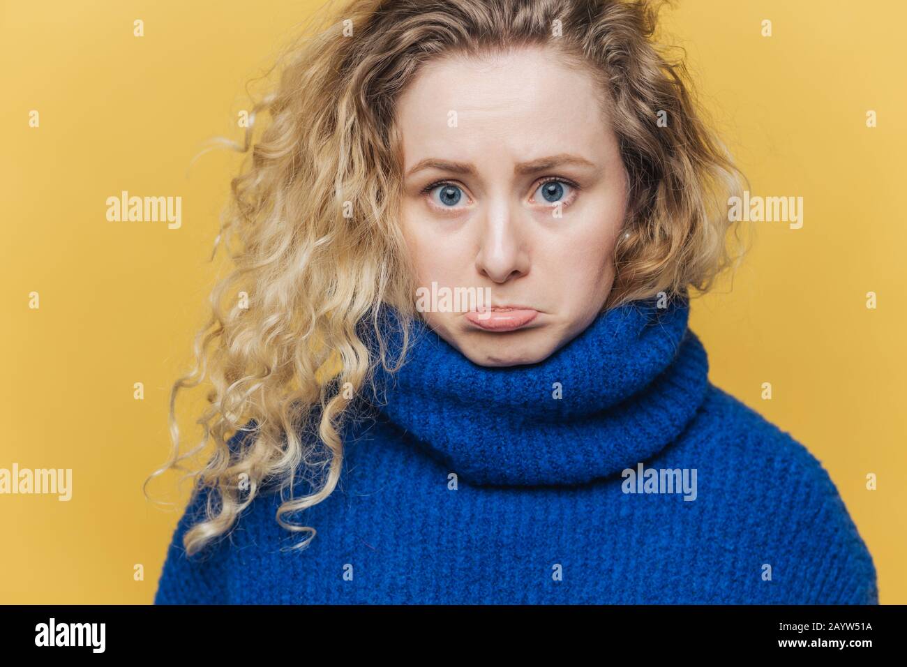 Attractive offended young female has curly bushy hair, curves lower lip in dissatisfaction, feels upset after quarrel with husband, wears blue sweater Stock Photo
