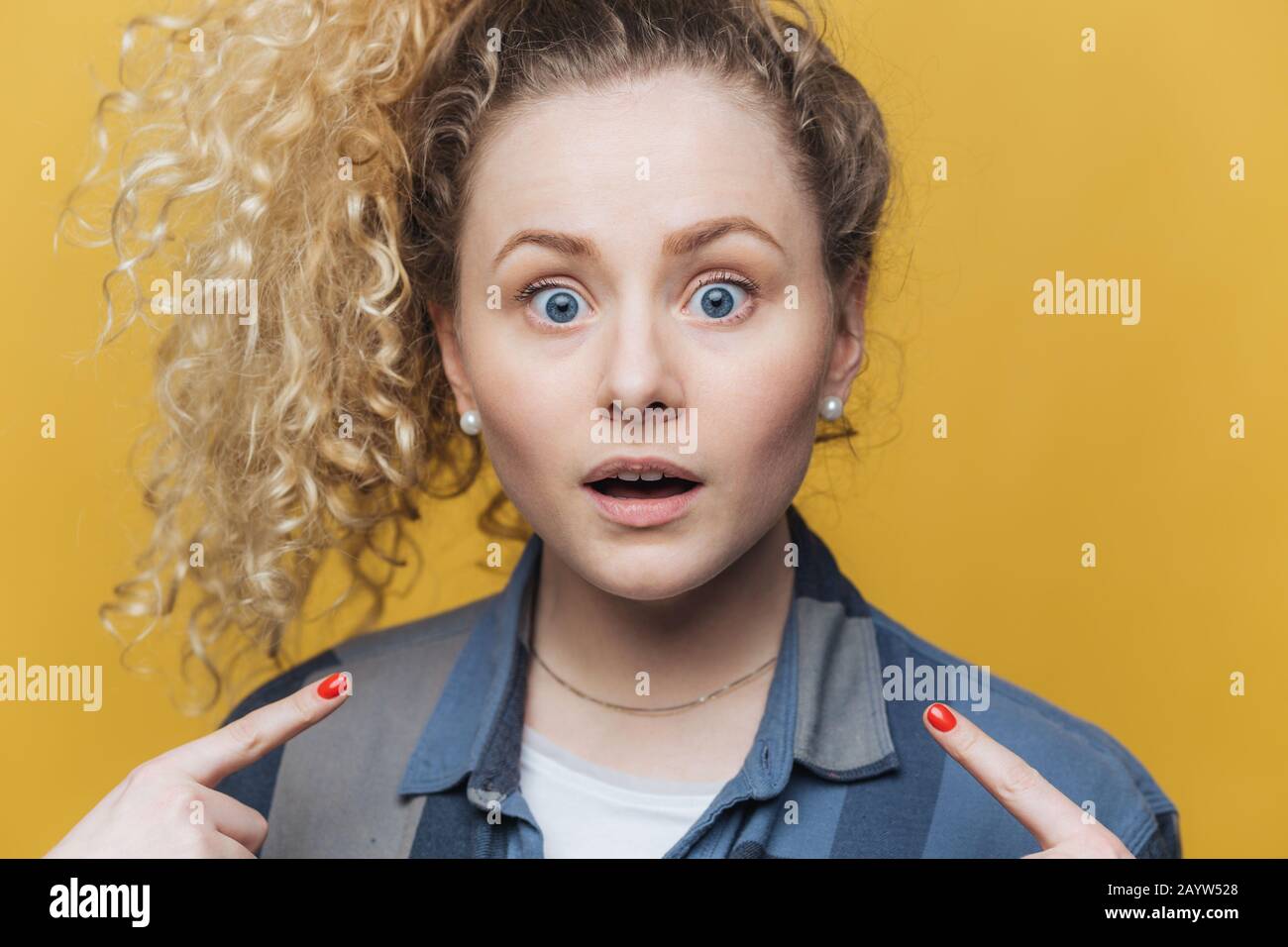 Horizontal shot of pleasant looking young curly woman indicates with fore fingers at herself, being shocked to recieve failure, dressed casually, has Stock Photo