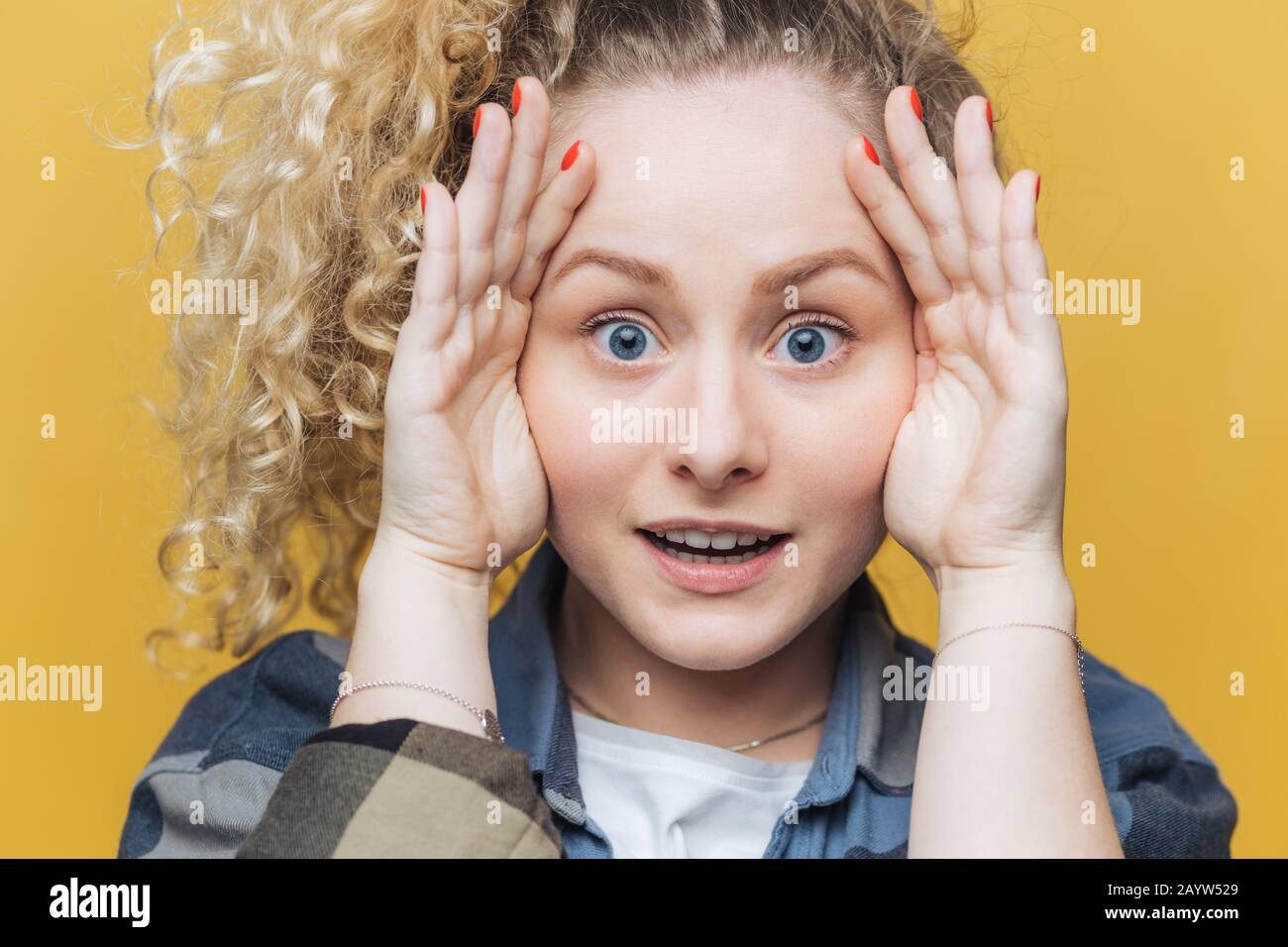 Close up shot of emotional cute female with healthy skin and blue eyes, keeps hands on head, looks scrupulously and surprisingly at camera, notices so Stock Photo