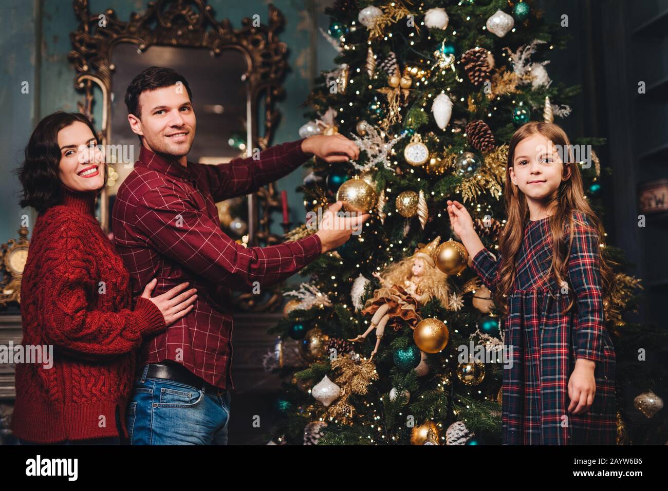 Friendly family of father, mother and daughter decorates New Year tree, being glad to have coming winter holidays, enjoy domestic atmosphere. Family o Stock Photo