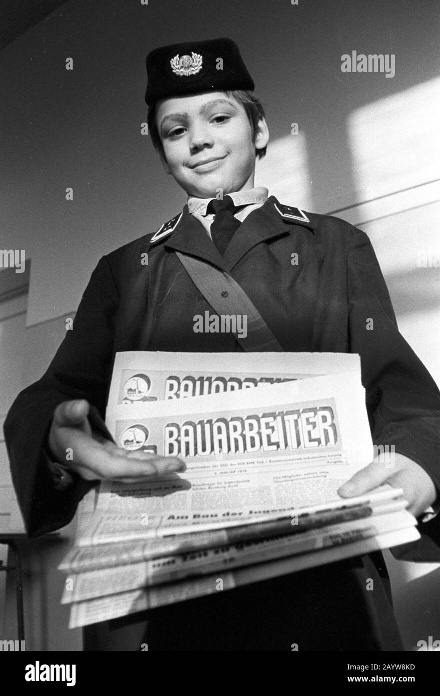 01 January 1979, Saxony, Leipzig: A child in a GDR postal uniform shows editions of the company newspaper "Der Bauarbeiter" at the end of the seventies. Larger combines and state-owned enterprises published their own newspapers - usually monthly or fortnightly - as company newspapers. This is the newspaper of the SED's operating party organisation of the VEB BMK Süd (construction and assembly combine), a combined company of Industriebau Leipzig. Photo: Volkmar Heinz/dpa-Zentralbild/ZB Stock Photo