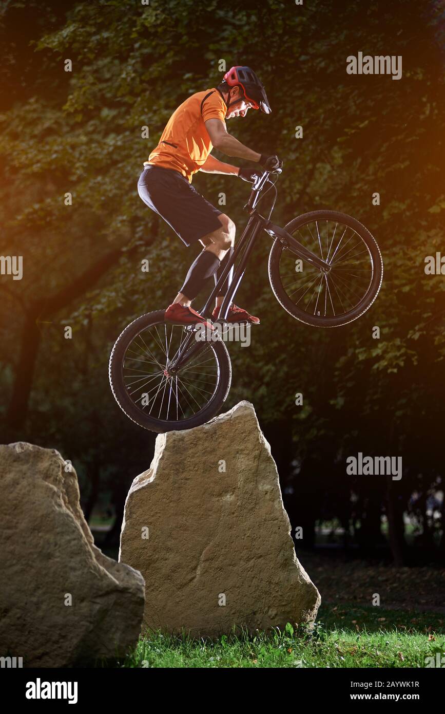 Extreme show of talented trail biker performing on big rocks standing on back wheel, in the evening in park, side view snapshot, concept of active lifestyle Stock Photo