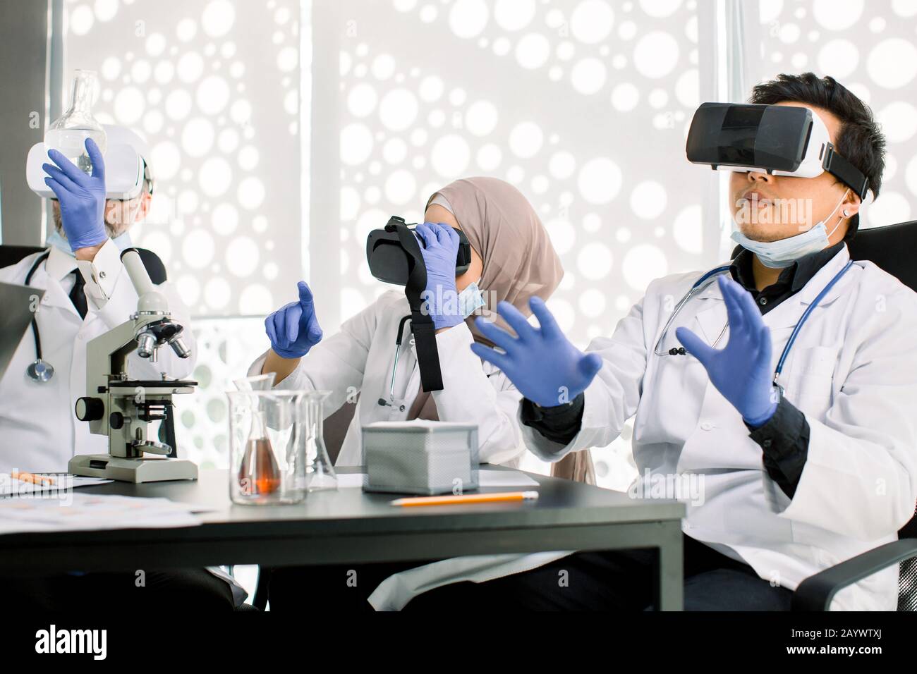 Team of scientists, Asian and Caucasian men, Muslim woman using virtual reality goggles for scientific research in laboratory, gesturing hands Stock Photo