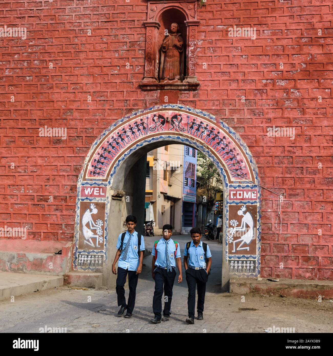 Three boys in school uniform walk on the road thatgoes through the old, red, Portuguese Zampa gateway in Diu Island. Stock Photo