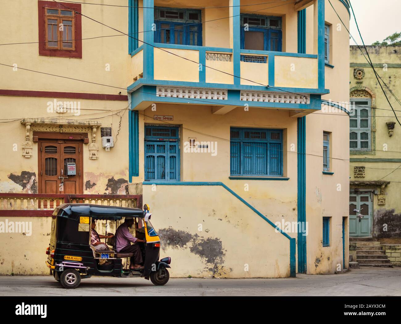 A quiet neighbourhood of old residential buildings with blue and brown windows in Diu. A rickshaw rides on the road. Stock Photo